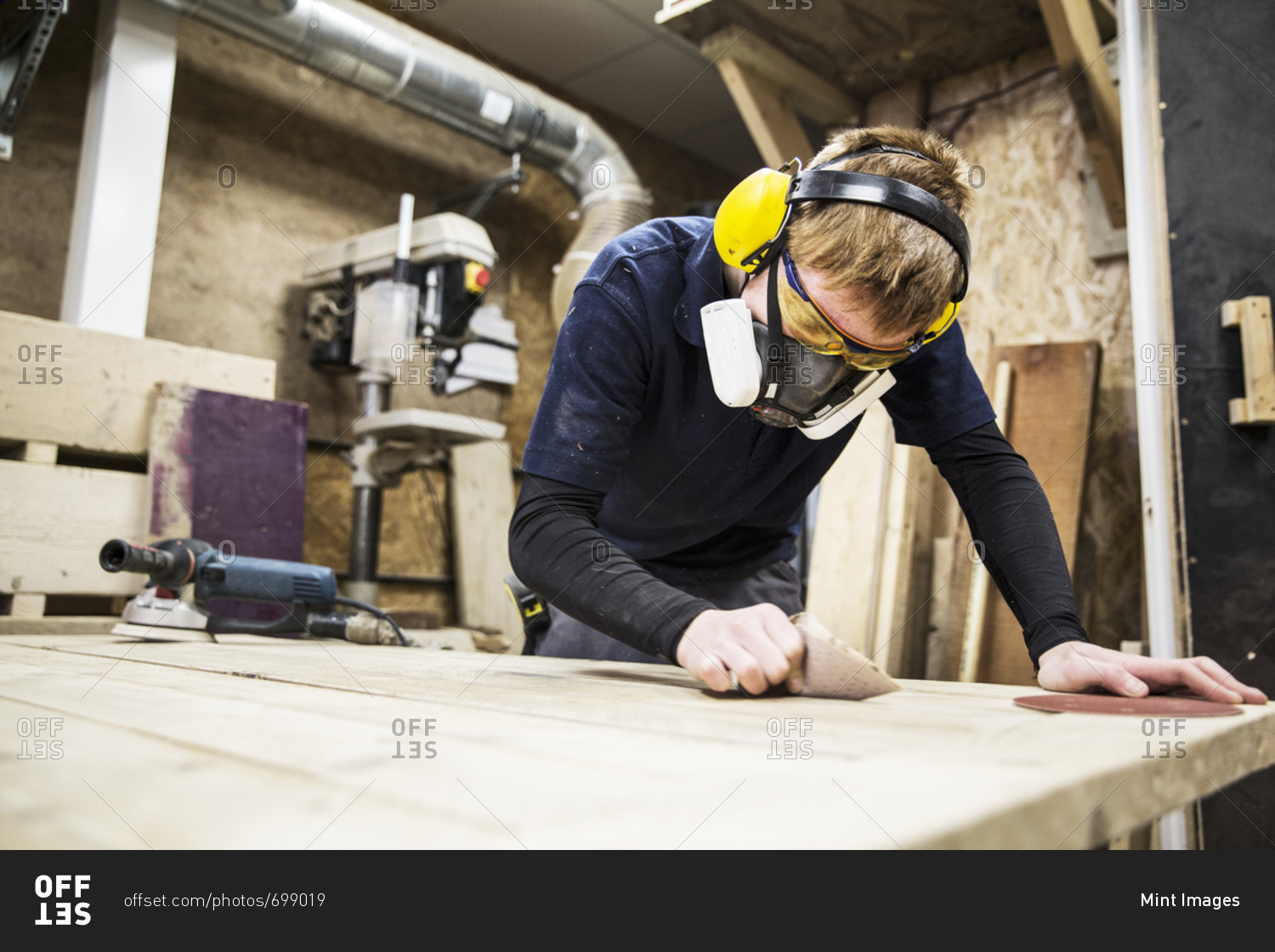 Man wearing ear protectors, protective goggles and dust mask standing in a warehouse, working on a piece of wood