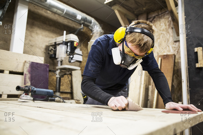 Man wearing ear protectors, protective goggles and dust mask standing in a warehouse, working on a piece of wood