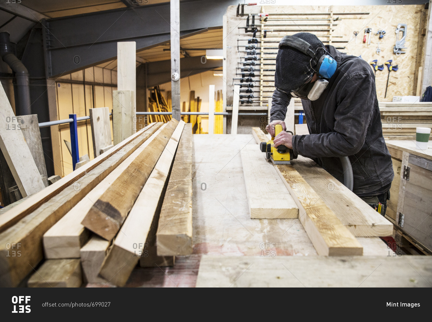 Man wearing ear protectors, protective goggles and dust mask standing in a warehouse, sanding planks of recycled wood