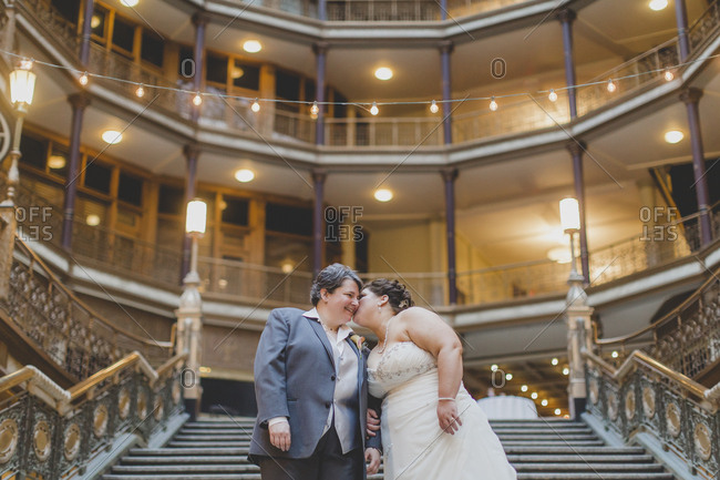 A lesbian couple share a tender kiss and smile on a grand staircase before entering their wedding reception