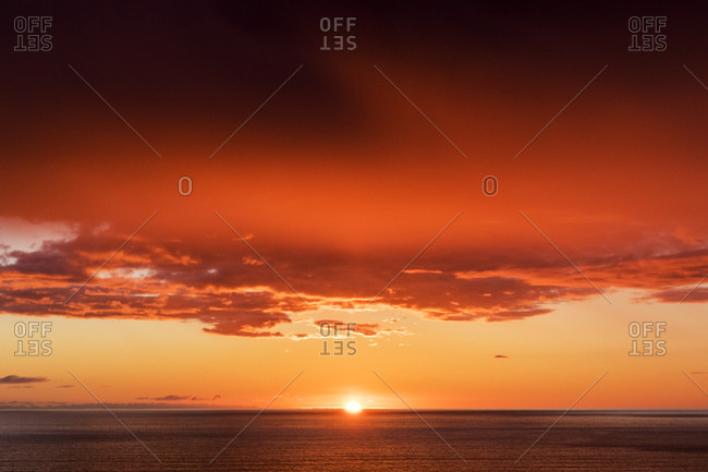 Bright orange sun dropping into the ocean as clouds glow