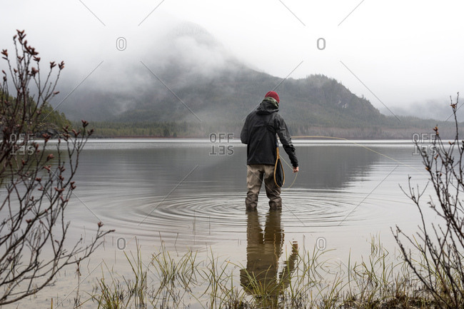 Rearview of fly fisherman standing in still waters of lake with mist shrouding hill side in distance