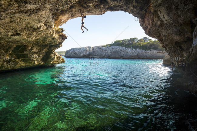 Adventurous rock climber climbing challenging route across natural arch, Cala Varques, Manacor, Mallorca, Spain