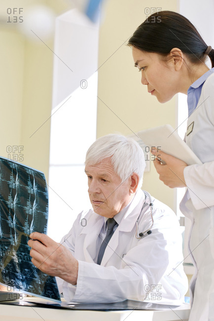 Mature male physician looking at radiograph of patient after MRI scan and analyzing his health conditions with young female assistant