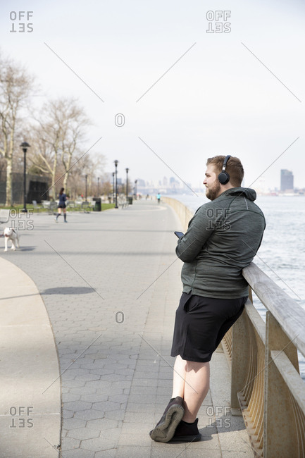 Full length of overweight man listening music on headphones while standing by river in city