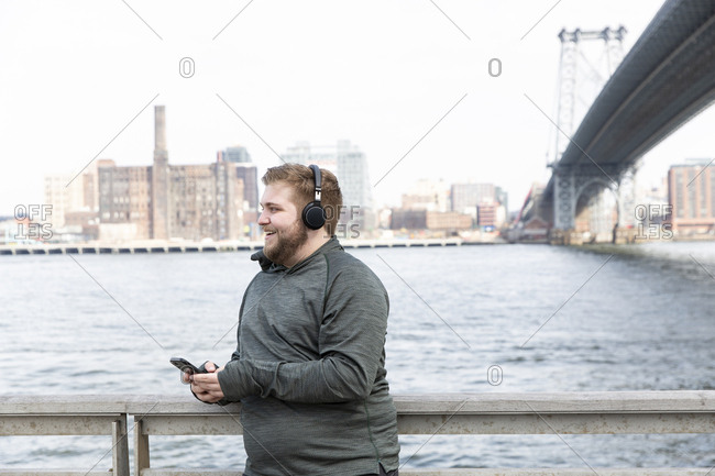 Smiling overweight man listening music on headphones while standing by river against Williamsburg Bridge in city