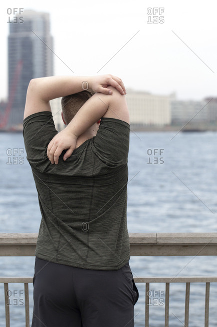 Rear view of overweight man stretching arms behind back while standing on bridge by river in city