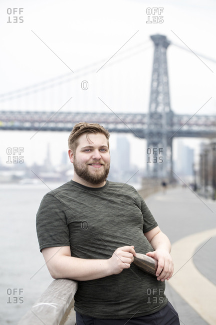 Portrait of smiling overweight man holding water bottle while standing against Williamsburg Bridge in city