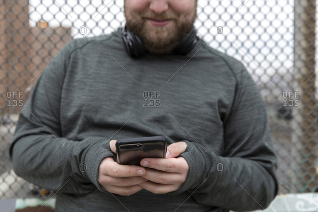 Midsection of overweight man using smart phone while standing against fence in city