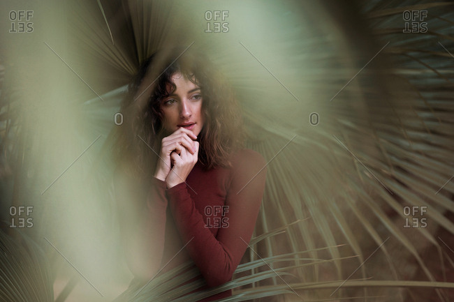 Beautiful sensual brunette with curls wearing casual dress and standing with hands on chest looking away among palm leaves