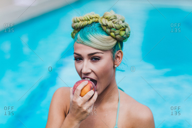 Swimsuit model with surreal hairstyle eating peach in front of pool