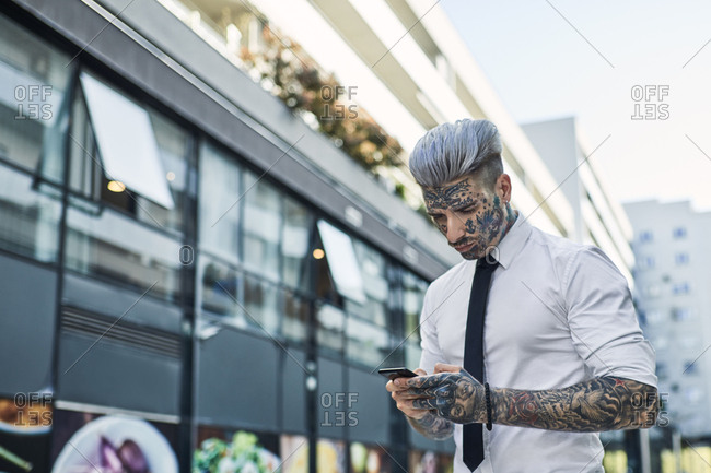Young businessman with tattooed face using smartphone stock photo - OFFSET
