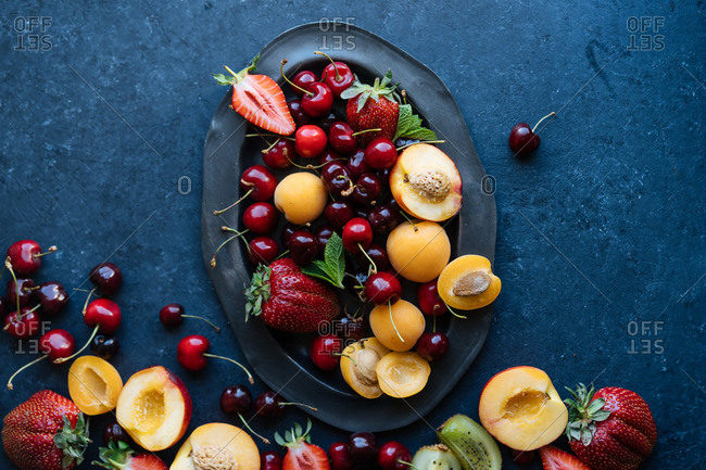 Top down view of selection of fresh summer fruits and berries overflowing from platter onto dark table top