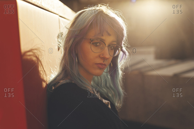 Portrait of confident young woman with blue hair and piercings leaning on red background and lens flare