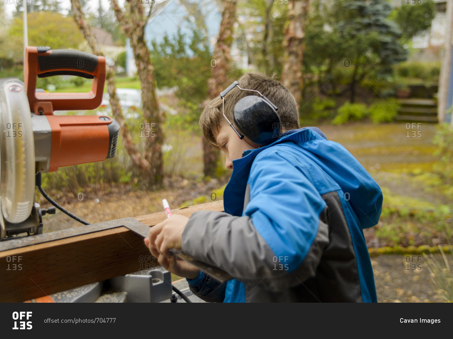 Boy wearing ear protectors while measuring wooden plank at backyard