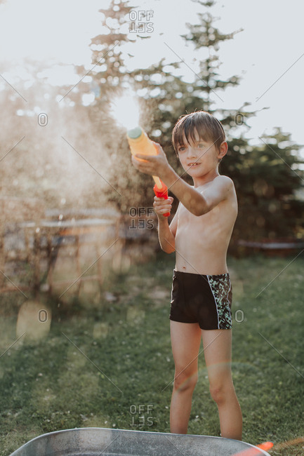 A portrait of a child wearing swimwear playing with a water gun on a hot summer day. A boy spraying water from a water pistol in a garden at summertime.