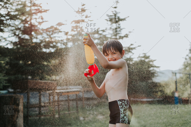 A portrait of an excited child spraying water all over himself on a hot summer day. A happy boy splashing with a water gun in a garden in the summertime.