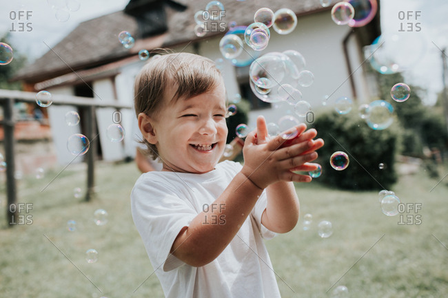 A happy child popping soap bubbles. A portrait of an excited toddler popping soap bubbles outside in a garden on a summer day.
