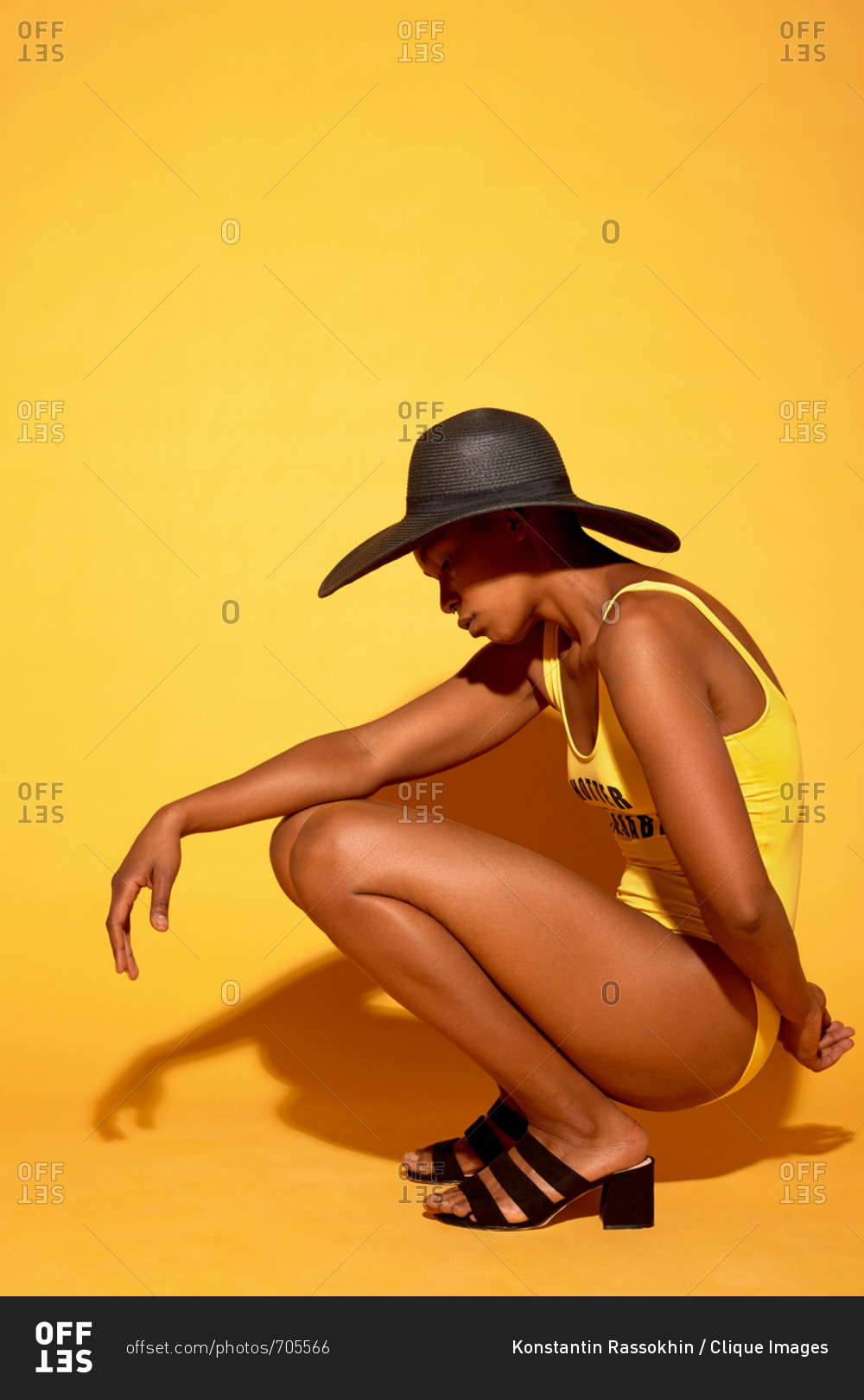 Fashion portrait of slim Black woman in one-piece swimsuit and sun hat posing on camera in squat position against orange background
