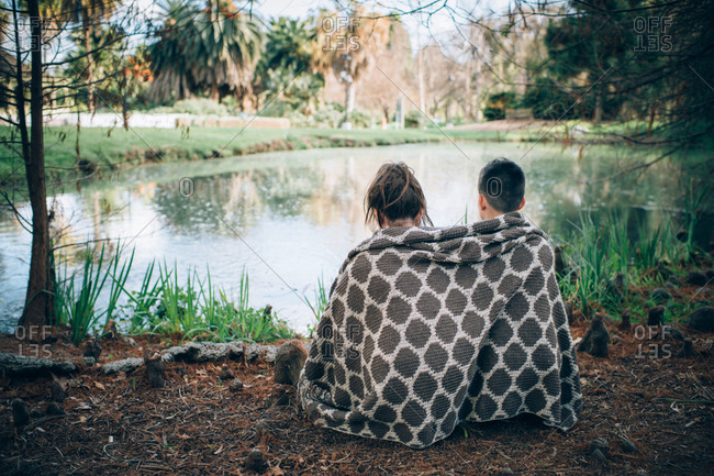 Siblings sitting in front of pond sharing thick woven blanket
