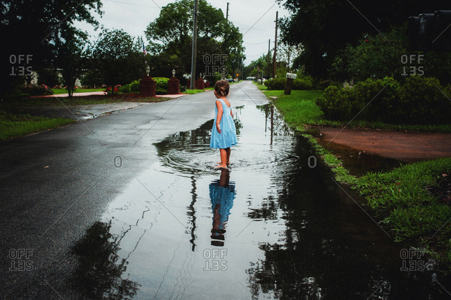Rearview of little girl in sun dress wading in puddle outside house