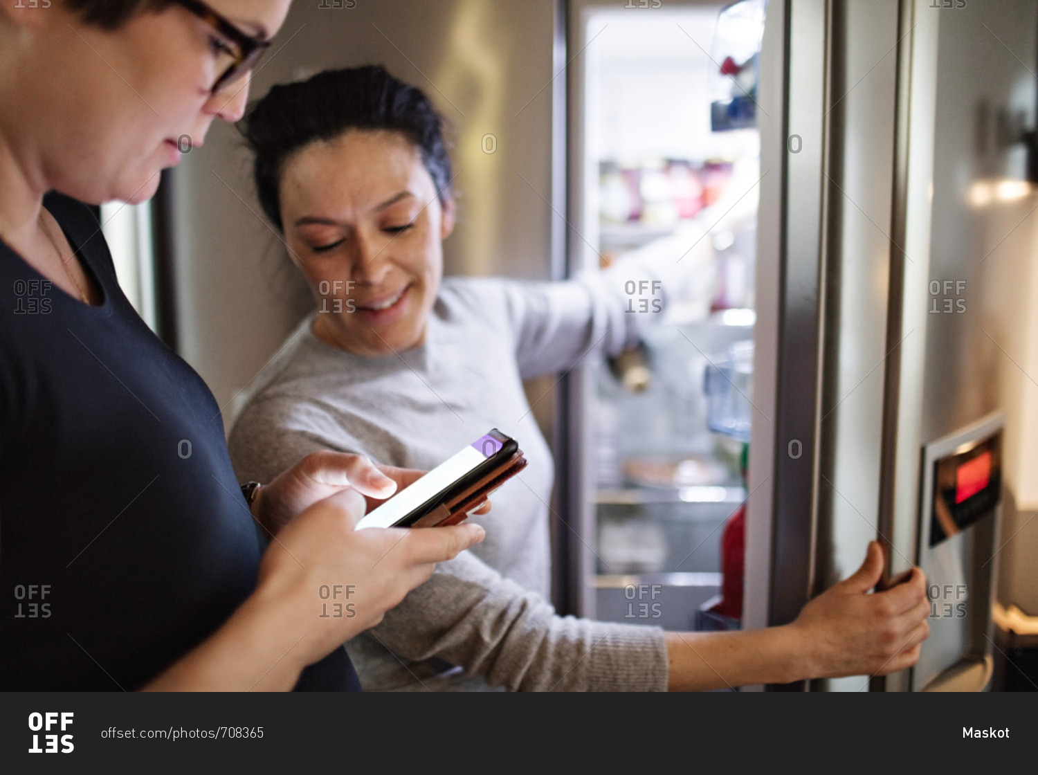 Woman showing mobile phone while girlfriend opening refrigerator in kitchen