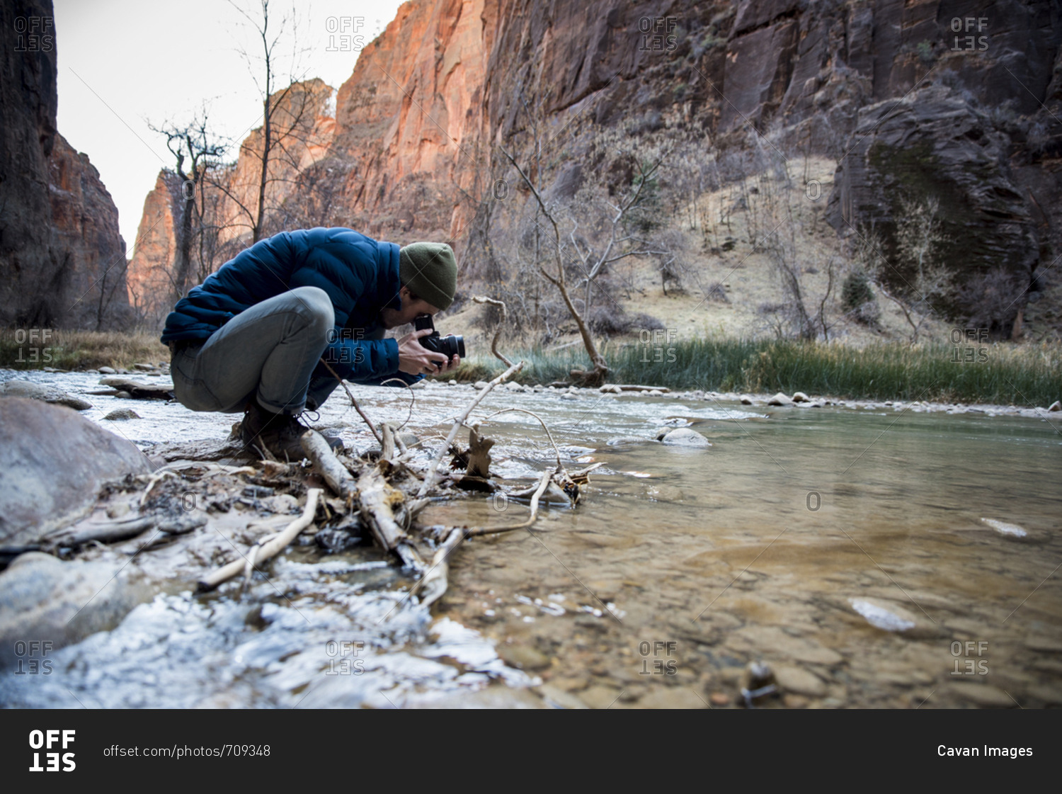 Side view of man photographing with camera while crouching by stream against rock formations