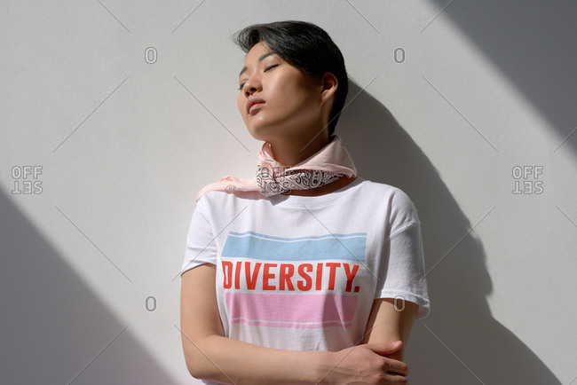 Portrait of attractive Asian girl in t-shirt with lettering and kerchief leaning on white wall with her eyes closed