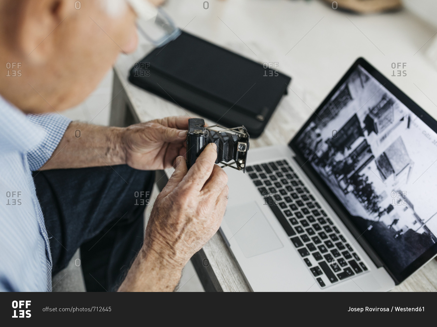 Senior man using laptop and holding his old photo camera- screen with old photo