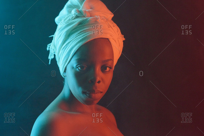 Waist-up portrait of young beautiful African American woman in white turban with bare shoulders looking at camera on dark background