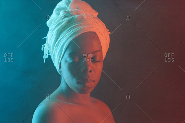 Waist-up portrait of young beautiful African woman in traditional head wrap posing in fog with bare shoulders and closed eyes on dark background