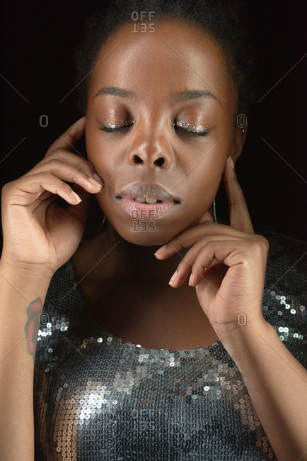 Waist-up portrait of young pretty African American woman in sequin top posing with glitter eye shadow makeup on dark background, her eyes closed