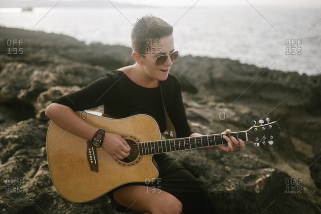 Young woman with short hair playing guitar at rocky beach