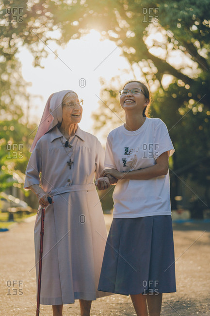 PHILIPPINES, APRIL 16, 2018: Young smiling female holding hand of cute elderly nun with stick on sunlit alley