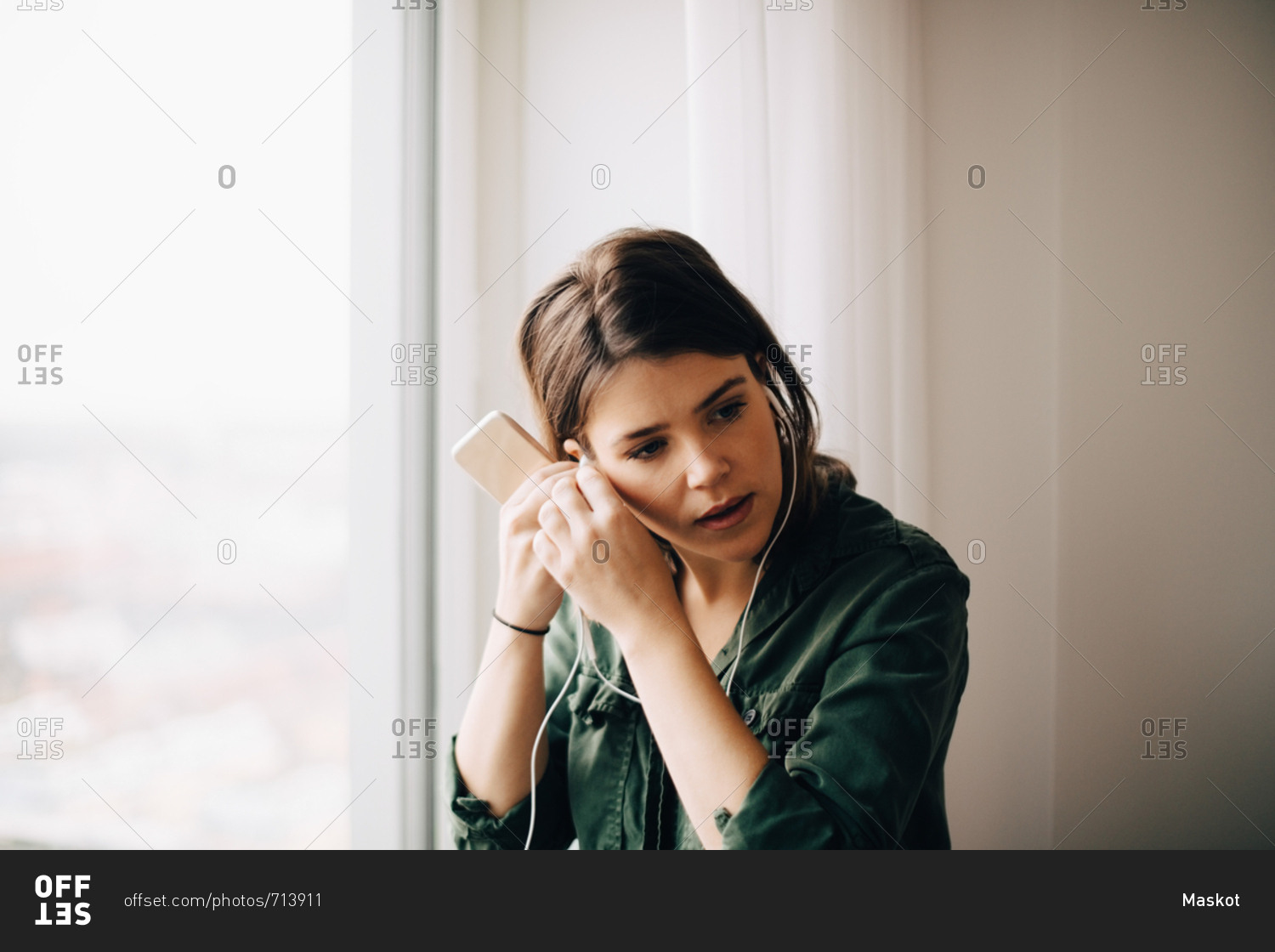 Businesswoman adjusting in-ear headphones while holding smart phone by window at creative office