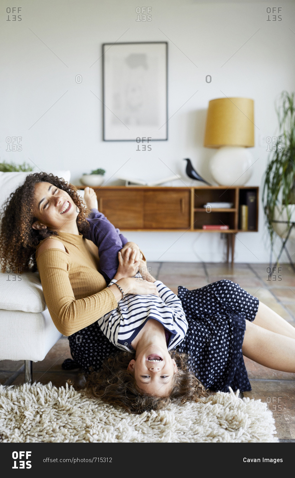 Happy mother holding daughter upside down while sitting on floor by sofa at home