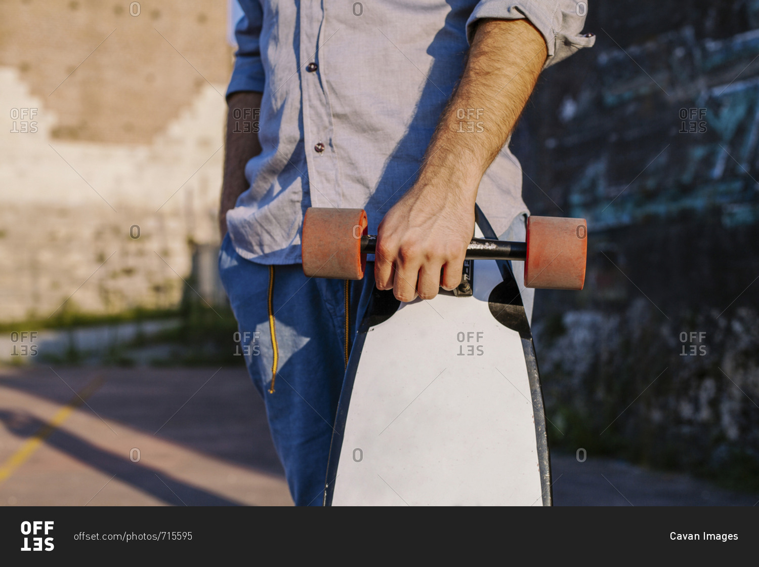 Midsection of man holding skateboard while standing against wall during sunset
