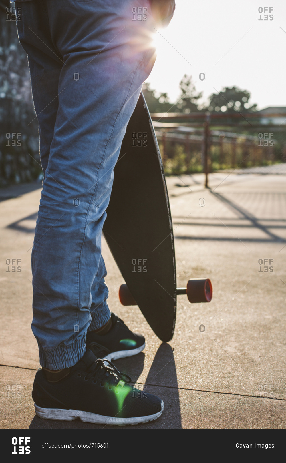 Low section of man holding skateboard while standing against sky during sunset