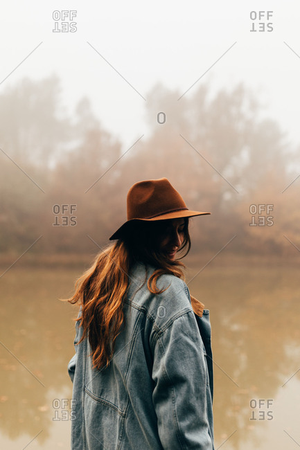 Pretty young woman in hat standing at small dirty pond in foggy day.
