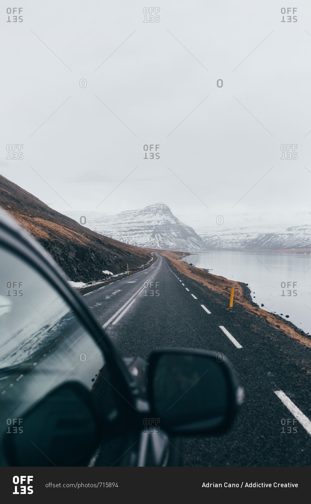 Black car driving down long road alongside cold lake with gloomy gray mountains on background in Iceland.