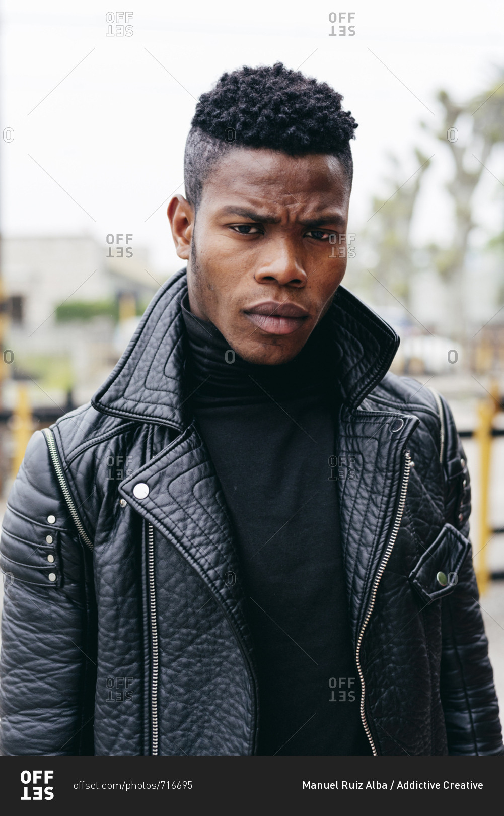 Serious african male in leather jacket raising hands behind head on blurred street background