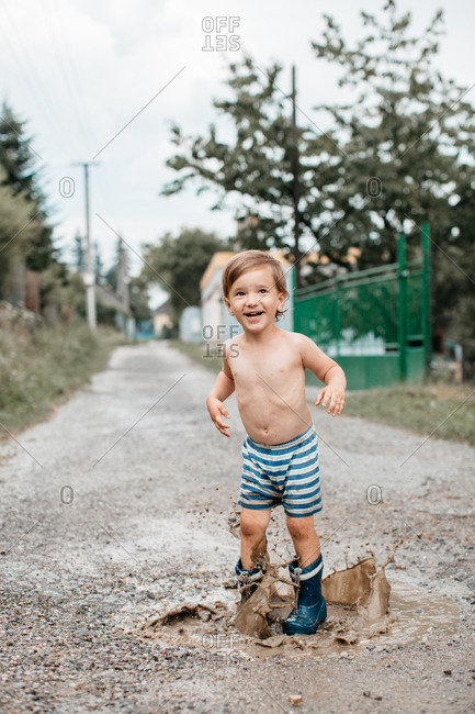 Portrait of a young smiling child in rain boots jumping puddles on a warm summer day. Happy toddler boy in wellington boots having fun splashing around on a road after the rain.