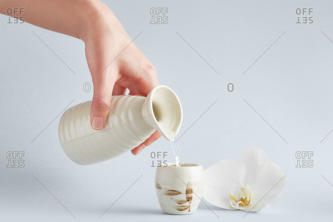 Crop view of hand holding white ceramic pitcher and pouring water into cup with floral ornament standing on white background with elegant white orchid nearby