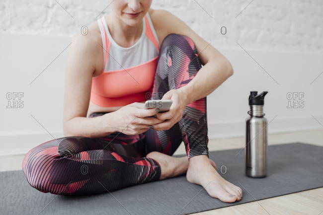 Cropped sportswoman sitting on mat and typing on cell phone