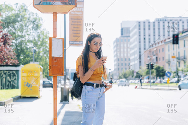 Young woman outdoors waiting at bus stop using smart phone