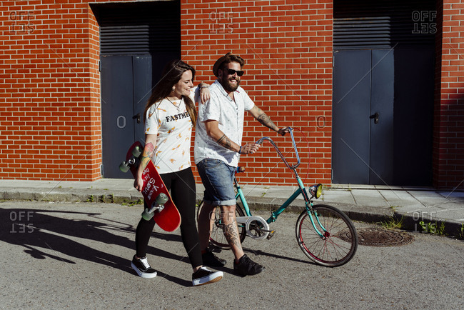 Trendy couple with tattoos walking on the street with a skateboard and a vintage bicycle