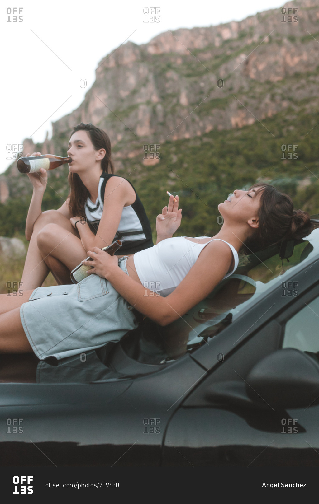 Side view of two girls sitting on the car hood smoking and drinking a