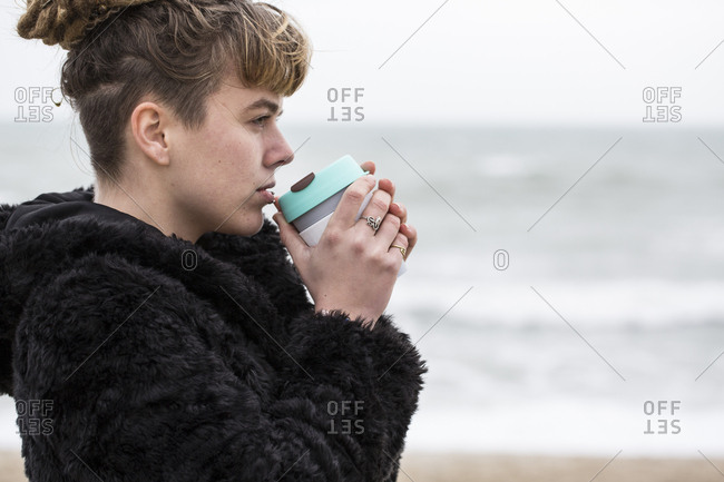 Portrait of young woman with brown hair and dreadlocks and a lip piercing wearing black furry jacket, standing by ocean, holding mug to her lips