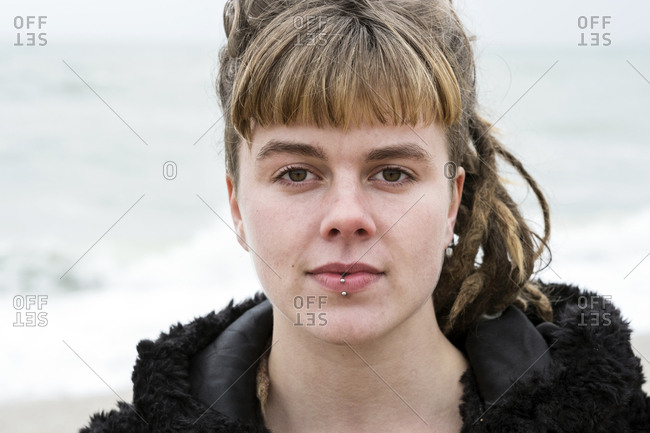 Portrait of young woman with brown hair and dreadlocks and a lip piercing wearing black furry jacket, looking at camera