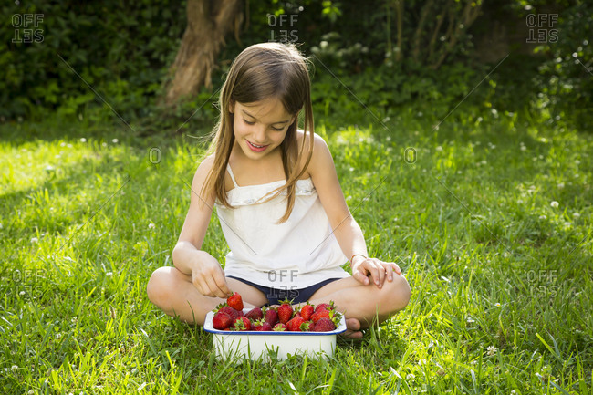 Smiling little girl sitting on meadow with bowl of strawberries